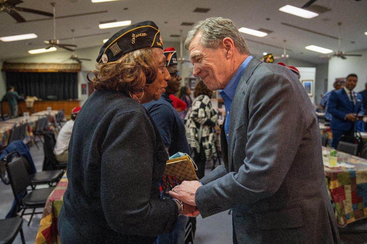 Roy shakes a veteran's hand at an event for Veterans Day. He wears a gray suit, and the veteran whose hand he is shaking wears a jacket and a veteran military cap.