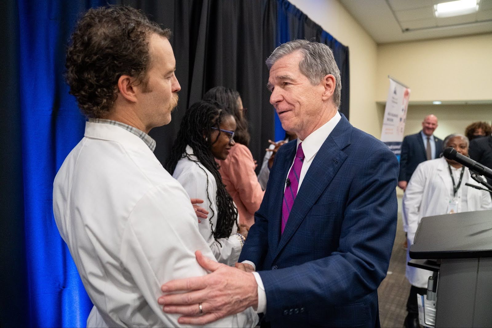 Roy speaks with a person in a white coat at an event supporting NC's Medicaid expansion. He wears a suit and tie.