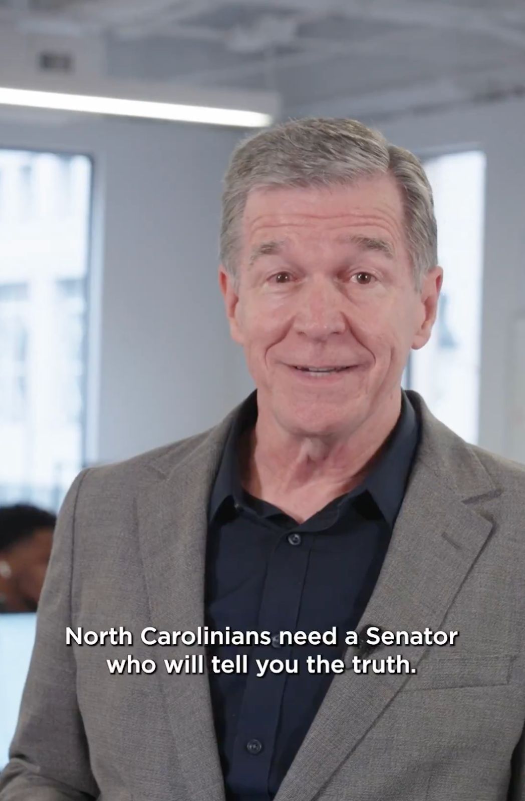 Roy wears a button-down shirt and a suit jacket at campaign headquarters and speaks to the camera. Subtitles read: “North Carolinians need a Senator who will tell you the truth.”