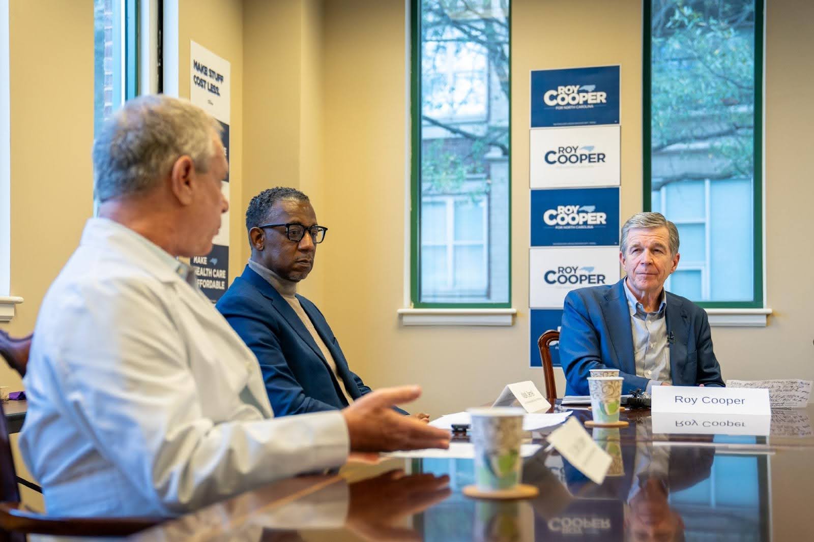 Roy participating in a roundtable discussion about health care. He wears a button-down shirt and a blue blazer, and is seated with two other people.